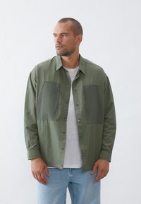 Green shirt jacket with two large chest pockets, button closure, and a lightweight texture; worn over a white t-shirt with blue jeans.