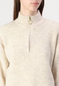 Beige ribbet genser med høy krage, med halv-glidelås og sirkulært gullhåndtak. Myk tekstur og jevn mønster gjennom hele.