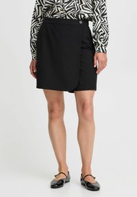 Zwarte wikkelrok met een gladde textuur, voorzien van een sluiting met zijknop. Gecombineerd met een gebloemd blouse en zwarte flats.