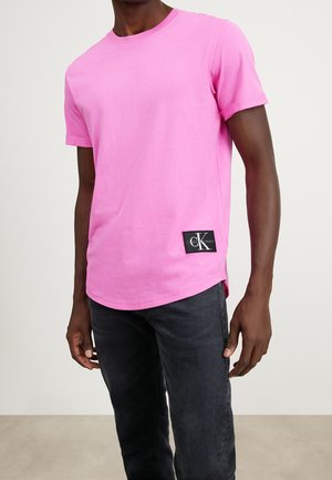 Rosa Baumwoll-T-Shirt mit Rundhalsausschnitt, kurzen umgeschlagenen Ärmeln und einem Calvin Klein-Logo-Patch am Saum. Kombiniert mit dunkelblauen Jeans.