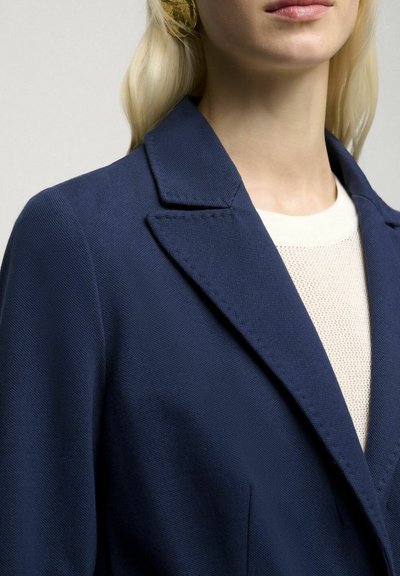 Marineblauwe textuurblazer met notch-lapels, opvallend displayed kraag en één zichtbare zak. Lichtgekleurde trui eronder.