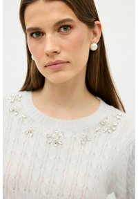 Vaaleanharmaa neulottu villapaita, jossa kukkakoristeisia helmiä pääntiellä, ja jossa on resorikaulus sekä teksturoitu palmikkoneulekuvio.