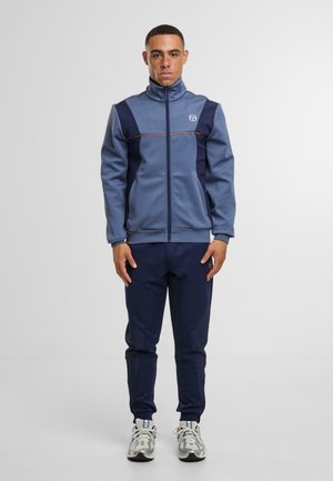 Pánský sportovní teplákový komplekt v modré barvě, obsahující zipovou bundu s námořnickými panely a oranžovými akcenty. Zúžené námořnické kalhoty, doplněné šedými teniskami.