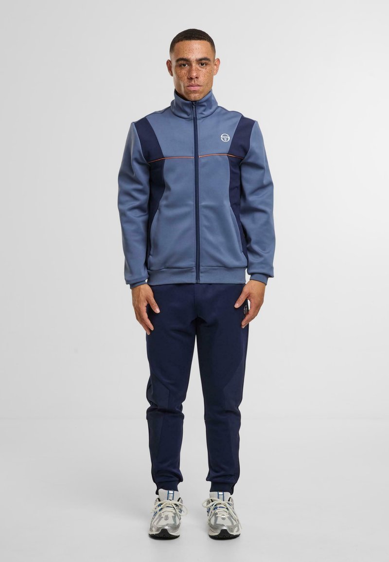 Pánský sportovní teplákový komplekt v modré barvě, obsahující zipovou bundu s námořnickými panely a oranžovými akcenty. Zúžené námořnické kalhoty, doplněné šedými teniskami.