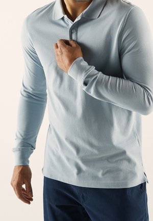 Hellblaues langärmeliges Poloshirt mit Kragen, zwei Knöpfen an der Knopfleiste und seitlichen Schlitzen, hergestellt aus einem weichen, strukturierten Stoff. Kombiniert mit dunkler Hose.