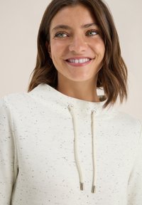 Ljus krämfärgad hoodie med ett fläckigt mönster, som har en dragsko i huvan och silverf�ärgade detaljer. Den mjuka texturen fulländar designen.