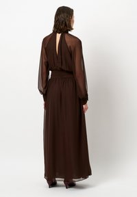 Bruine chiffon maxi-jurk met lange mouwen, open rugdetail en gathers op de taille. Soepele textuur, vloeiende silhouet en met knoopjes afgewerkte manchetten.