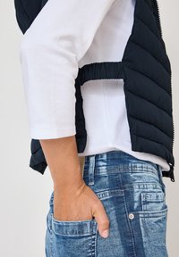 Gepolsterte marineblaue Weste mit hohem Kragen, ausgestattet mit einem Reißverschluss und seitlichen Ausschnitten, getragen über einem weißen Langarmshirt und hellblauen Jeans.