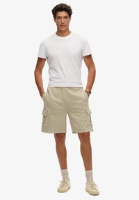 Witte crewneck t-shirt gecombineerd met beige cargoshorts met twee zijzakken en een relaxte pasvorm. Sneakers in neutrale kleuren maken de outfit compleet.