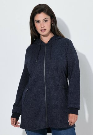 Vrouw draagt een donkerblauwe getextureerde capuchontrui met rits en zijzakken, staand tegen een effen lichte achtergrond.