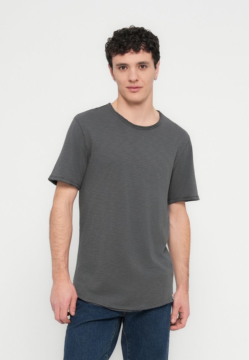 Young man with short curly hair wearing a plain dark gray short-sleeve t-shirt and blue jeans, standing against a light background.