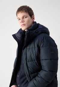 Doudoune bleu marine avec un col haut, fermeture éclair et patch de marque sur la manche. Présente une texture matelassée et une capuche amovible.