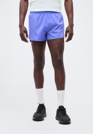 Mand iført hvide sokker, sorte løbesko og lilla sportsshorts med Nike-logo mod ensfarvet baggrund.