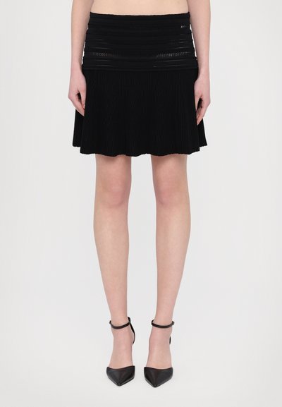 Mini-jupe plissée noire avec une taille structurée, tissu texturé et ourlet festonné, associée à des escarpins noirs à talons pointus.