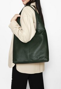 Donkergroene leren draagtas met een gestructureerde oppervlakte, slungelige vorm en brede banden, gedragen over een beige blouse. Geschikt voor casual gebruik.