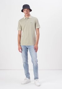 Lys grønn polo skjorte med en liten logo, kombinert med lyseblå jeans og hvite joggesko. Modellen har på seg en svart bøttehatt.