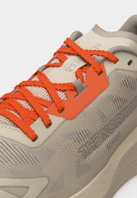 Μπεζ αθλητικό παπούτσι North Face με φωτεινά πορτοκαλί κορδόνια που περνούν μέσα από ξεχωριστό πορτοκαλί πάνελ οπών σε υφή επάνω μέρος.