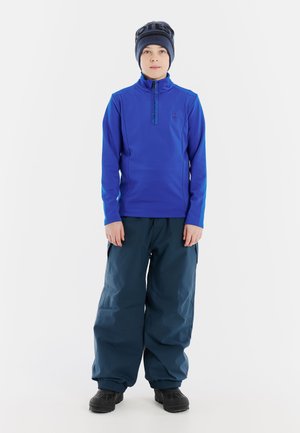 Maglia a maniche lunghe blu con collo a zip e pantaloni da neve blu scuro. L'outfit presenta una vestibilità comoda e stivaletti neri, adatto per attività invernali.