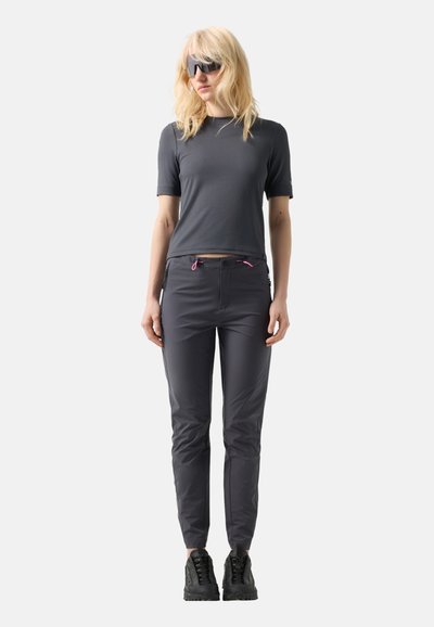 Blondýna s černými slunečními brýlemi, tmavě šedým tričkem s krátkým rukávem, úzkými tmavě šedými kalhotami s růžovými šňůrkami a černými teniskami.