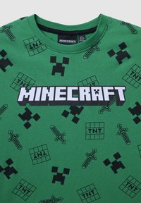 Maglietta verde in cotone con un motivo ripetuto a tema Minecraft, comprendente spade pixelate e disegni di creeper, con la scritta audace "MINECRAFT".