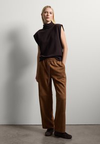 Haut à col roulé sans manches marron foncé, associé à un pantalon marron ample à taille haute avec un cordon de serrage, et des chaussures marron foncé.