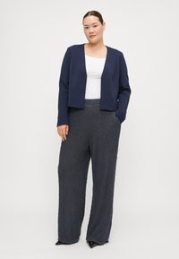 Veste cropped marine en tissu lisse, associée à un pantalon large plissé gris foncé. La tenue se compose d'un simple top blanc.