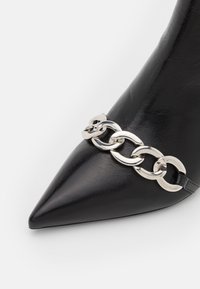 Mustat nahkaiset korkokengät, joissa on terävä nenä ja hopeaketju korostus edessä, korostaen sileää tekstuuria ja sulavalinjaista muotoilua.