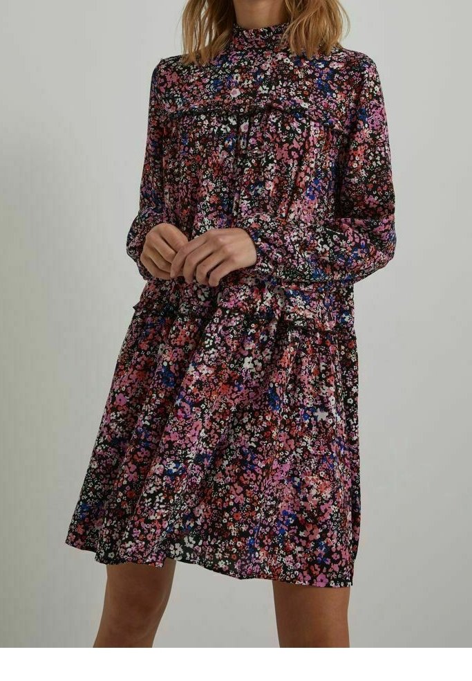Femme portant une robe à manches longues et col montant avec un imprimé floral rose, violet, blanc et bleu, debout devant un fond uni.