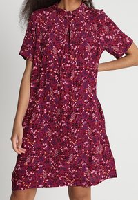 Kortærmet kjole i mørkerødt stof med blomstermønster i pink og lilla nuancer, med krave og knapdetalje ved halsen.