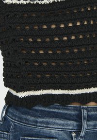 Fekete horgolt felső, nyitott szövésű dizájnnal, amelynek scalloped fehér szegélye van. Kék farmernadrággal párosítva, amely a laza stílust mutatja.