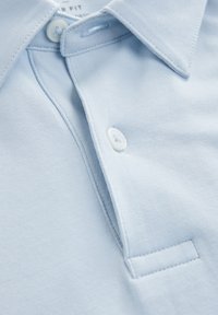 Šviesiai mėlynas polo marškinėliai su minkšta tekstūra, turintys tradicinį apykaklę, mygtukų užtrauktuką ir mažą siuvinėtą kišenės detalę.