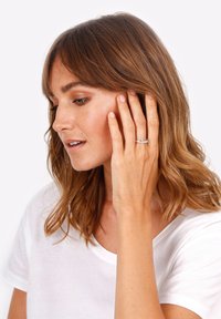 Anello d'argento con bande intrecciate, caratterizzato da piccole pietre trasparenti incastonate in una base metallica lucida, esposto sulla mano di un modello.
