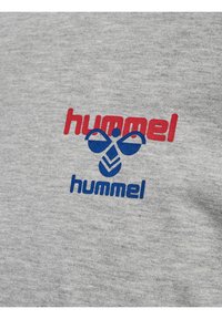 Czerwono-niebieskie logo marki „hummel” z stylizowaną grafiką pszczoły na jasnoszarym materiale.