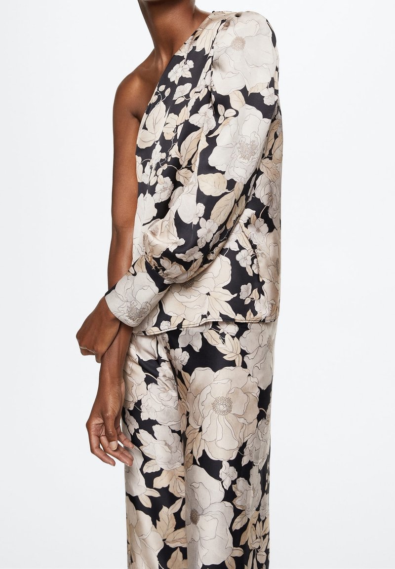 Personne portant un costume floral asymétrique avec de grandes fleurs beige et blanches sur fond noir, debout les bras croisés.