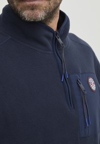 Marineblauer Fleece-Pullover mit hohem Kragen, Halb-Reißverschluss und einer Brusttasche mit rundem Logo. Blaue Reißverschlussanhänger und weiche Textur.