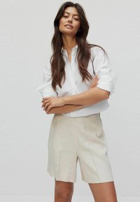 Witte overhemd met opgerolde mouwen, gecombineerd met lichtbeige shorts. De shorts hebben een rechte snit en subtiele plooien.