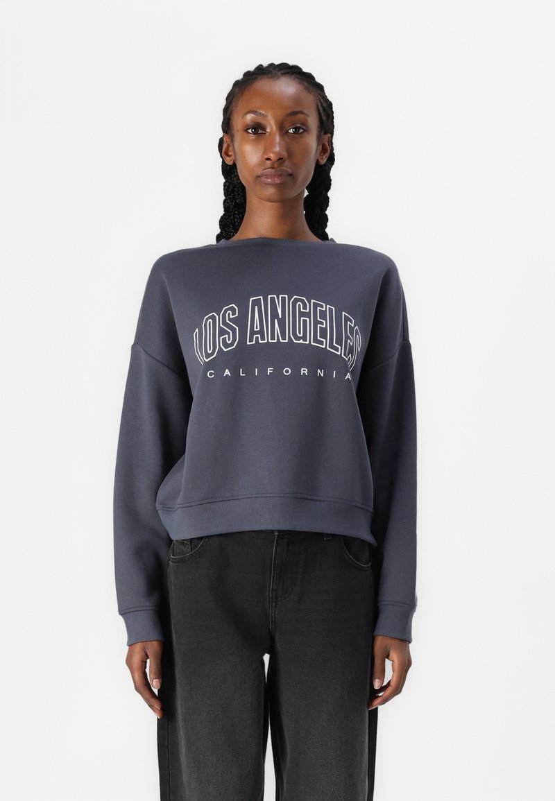 Kvinde med flettet hår iført en mørkegrå sweatshirt med teksten "LOS ANGELES CALIFORNIA" og sorte bukser, stående mod en hvid baggrund.