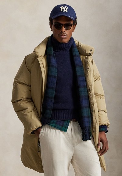 Tan puffert jakke, navyblå sweater, ternet tørklæde med grøn og blå, creme farvede kordfløjlsbukser, blå baseballkasket, mørke solbriller.
