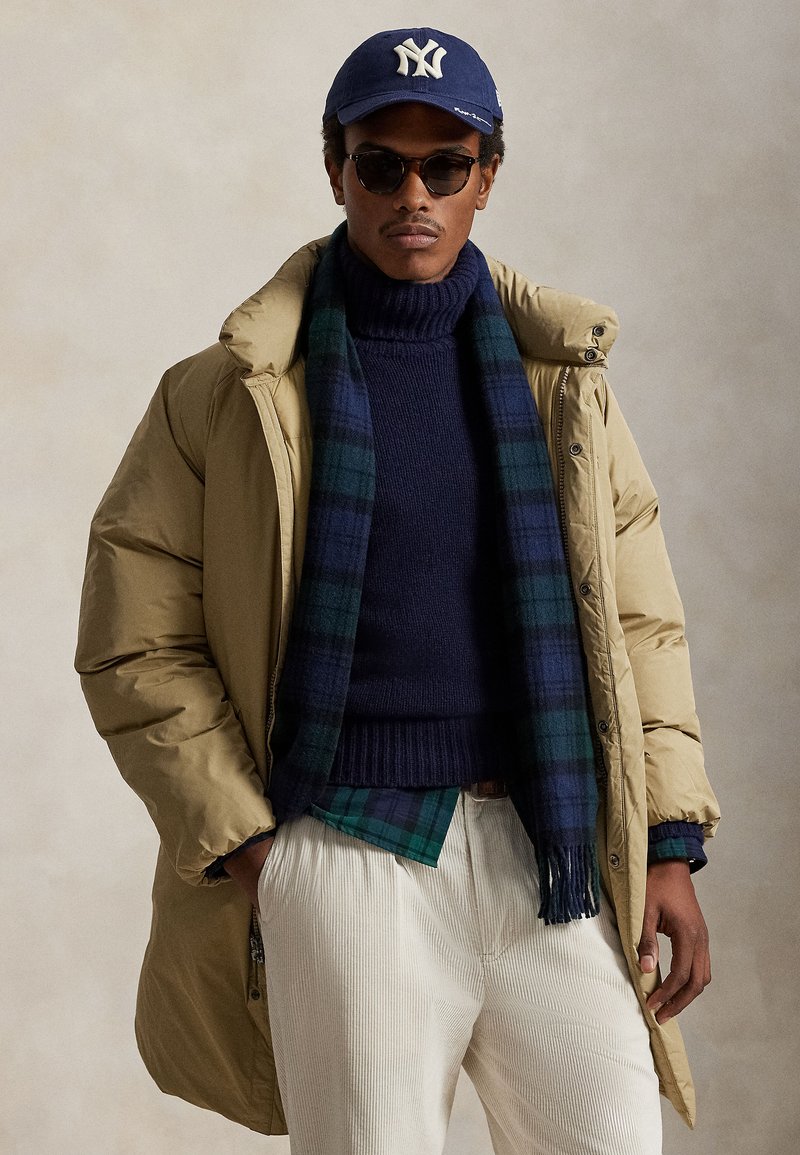 Bēšs pūkains mētelis, tumši zilas krāsas džemperis, pledveida šalle zaļā un zilā krāsā, krēmkrāsas korduroja bikses, zila beisbola cepure, tumši saulesbrilles.