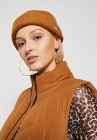 Brun ribbet strikket lue kombinert med en beige ermeløs puffer vest over en leopardmønstret langermet topp. Store hoops øreringer synlige.