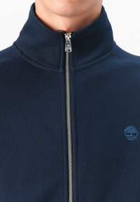 Jachetă bleumarin cu fermoar, având un guler înalt, textură ribbed și un mic logo al unui copac albastru pe partea stângă a pieptului.