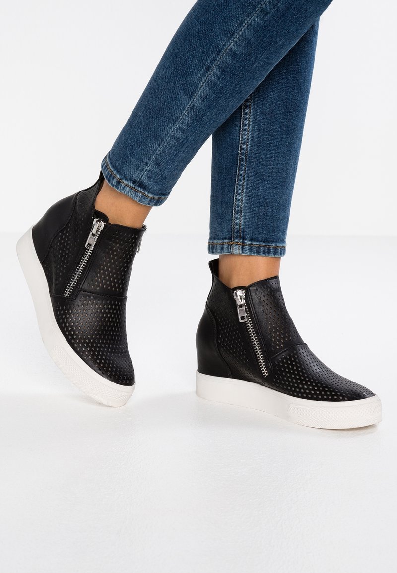 Botines de tobillo negros con perforaciones, con tacón de cuña y suela blanca, que presentan un cierre de cremallera lateral y detalles de cuero suave.