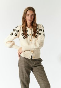 Cardigan lavorato a maglia crema con maniche a sbuffo testurizzate, adornato da ricami floreali neri e dorati. Presenta bottoni dorati lungo la parte anteriore.
