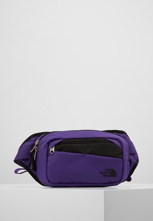 Lilla og sort North Face bæltetaske med flere lynlåslommer, placeret på en hvid overflade mod en ensfarvet baggrund.