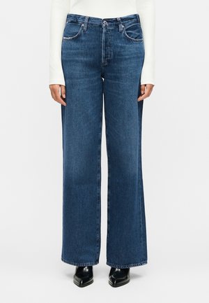 Μπροστινή όψη ατόμου που φορά ψηλόμεσα, φαρδιά σκουρομπλε τζιν με μαύρα μυτερά παπούτσια και λευκό μακρυμάνικο τοπ, με τα χέρια χαλαρά στο πλάι.