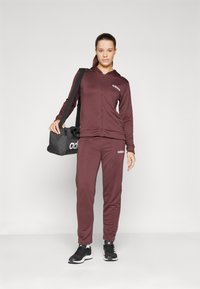 Maroon athletic tracksuit with a zip-up hoodie, matching pants, and black sports shoes. Features Adidas logo in white. Includes a black duffle bag.