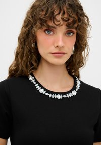 Schwarzes Kleid mit rundem Ausschnitt, verziert mit einer Reihe ovaler und marqueeförmiger klarer Strasssteine. Kurze Ärmel und glatte Stoffstruktur.