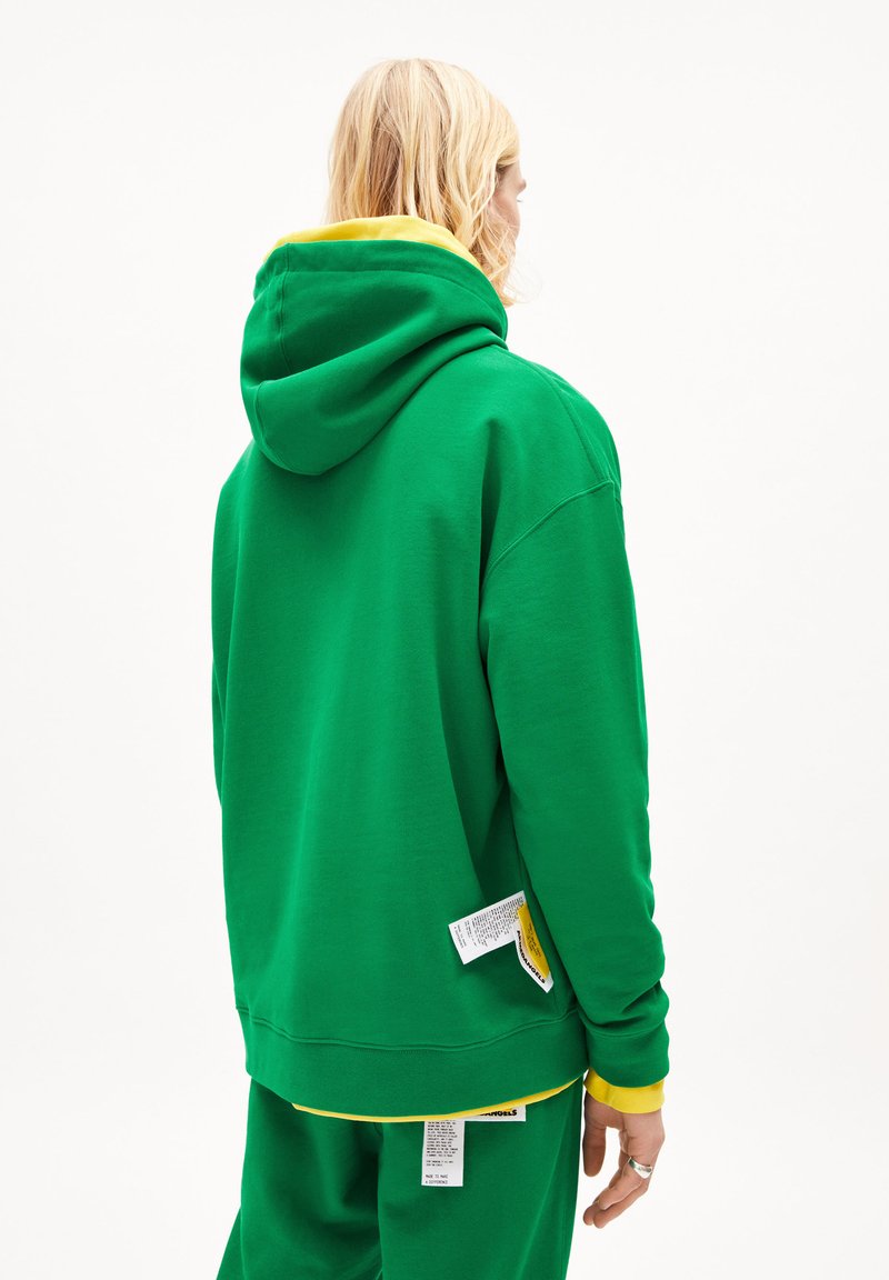 Person mit langen blonden Haaren, die einen grünen Hoodie und Hosen mit gelbem Innenfutter trägt, von hinten sichtbar, wobei die Etiketten auf der Kleidung zu sehen sind.