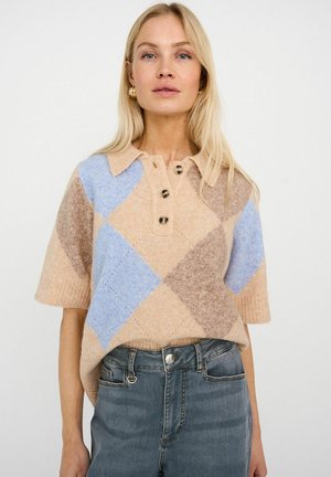Blondinka v bež, modrem in rjavem argyle pletenem polo majici s kratkimi rokavi in modrih kavbojkah z visokim pasom na belem ozadju.