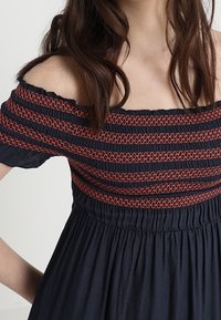 Kvinna i en marinblå off-shoulder-klänning med röda broderade horisontella ränder över bröstet och övre ärmar.